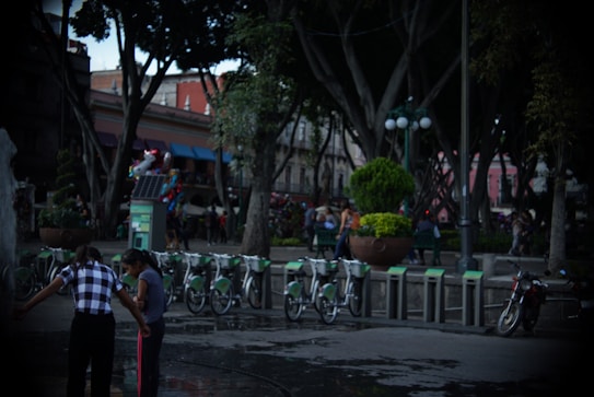 A public park setting with large trees and a bicycle rental station. People are enjoying the park, some gathered near a water fountain. A variety of colorful balloons can be seen, adding vibrancy to the scene. In the background, there are urban buildings and bright street lighting creating a lively atmosphere.