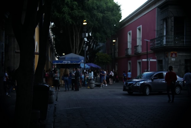 A dimly lit urban street scene with people gathering near an outdoor food stall under a blue canopy. The setting includes a tree-lined avenue and adjacent red and yellow buildings. Cars are parked and passing by on the road, contributing to a bustling atmosphere.