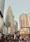 A bustling urban landscape featuring modern skyscrapers and city architecture, with various advertisements and signs visible on the buildings. The scene is lively with people walking in the foreground, surrounded by shops and commercial establishments.