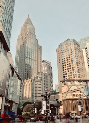 A bustling urban landscape featuring modern skyscrapers and city architecture, with various advertisements and signs visible on the buildings. The scene is lively with people walking in the foreground, surrounded by shops and commercial establishments.