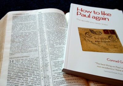 An open Bible is displayed with text visible, showing references to Corinthians. Beside it is a book titled 'How to like Paul again' by Conrad Gempf, which carries an image of an old letter with postal stamps. The arrangement suggests a reading or study setup.