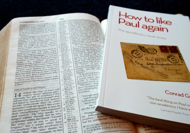 An open Bible is displayed with text visible, showing references to Corinthians. Beside it is a book titled 'How to like Paul again' by Conrad Gempf, which carries an image of an old letter with postal stamps. The arrangement suggests a reading or study setup.