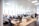 An open-plan office space with several people working at desks equipped with computers and other office supplies. The room has large windows allowing natural light to illuminate the space. The office is modern with white walls, ceiling lights, and wooden and white office furniture.