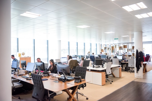 An open-plan office space with several people working at desks equipped with computers and other office supplies. The room has large windows allowing natural light to illuminate the space. The office is modern with white walls, ceiling lights, and wooden and white office furniture.