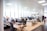 An open-plan office space with several people working at desks equipped with computers and other office supplies. The room has large windows allowing natural light to illuminate the space. The office is modern with white walls, ceiling lights, and wooden and white office furniture.