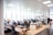 An open-plan office space with several people working at desks equipped with computers and other office supplies. The room has large windows allowing natural light to illuminate the space. The office is modern with white walls, ceiling lights, and wooden and white office furniture.