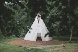 A white, conical tent resembling a teepee is situated in a clearing surrounded by dense green foliage. The tent is set on a circular patch of wood chips or mulch, with grass and trees in the background. A few tree branches frame the scene, enhancing the secluded, nature-centric atmosphere.