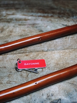 A red key tag labeled 'BAYONNE' attached to a single silver key lies on a rustic wooden surface. Two polished wooden rods or tools are positioned diagonally on either side.