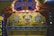 A carnival game booth featuring bright, colorful artwork with a large clown face at its center, surrounded by target designs. The booth is decorated with red curtains and lit by carnival lights providing a retro, playful atmosphere. Various cans and targets are part of the game setup.