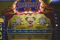 A carnival game booth featuring bright, colorful artwork with a large clown face at its center, surrounded by target designs. The booth is decorated with red curtains and lit by carnival lights providing a retro, playful atmosphere. Various cans and targets are part of the game setup.