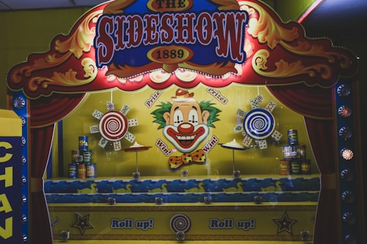A carnival game booth featuring bright, colorful artwork with a large clown face at its center, surrounded by target designs. The booth is decorated with red curtains and lit by carnival lights providing a retro, playful atmosphere. Various cans and targets are part of the game setup.