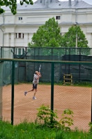 A person is playing tennis on a clay court, enclosed by a chain-link fence. The player is captured mid-motion, swinging a tennis racket. In the background, there is a large white building with classical architectural features and lush green trees.