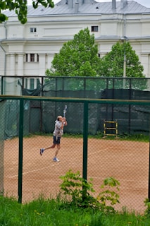 A person is playing tennis on a clay court, enclosed by a chain-link fence. The player is captured mid-motion, swinging a tennis racket. In the background, there is a large white building with classical architectural features and lush green trees.