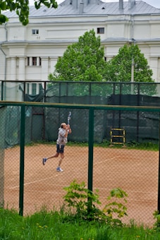 A person is playing tennis on a clay court, enclosed by a chain-link fence. The player is captured mid-motion, swinging a tennis racket. In the background, there is a large white building with classical architectural features and lush green trees.
