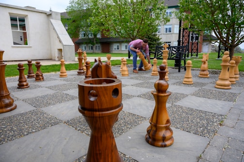 A person is engaged in moving a large wooden chess piece on an outdoor chessboard. The chess pieces are oversized and made of wood, situated on a concrete and stone-tiled ground. In the background, there are buildings and trees in a park-like setting, creating a serene and thoughtful atmosphere.