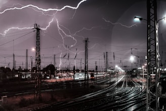 Lightning strikes illuminate a busy railway yard at night. Numerous overhead power lines and pylons are silhouetted against the dark sky, with multiple railway tracks converging in the foreground.