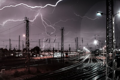 Lightning strikes illuminate a busy railway yard at night. Numerous overhead power lines and pylons are silhouetted against the dark sky, with multiple railway tracks converging in the foreground.