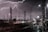 Lightning strikes illuminate a busy railway yard at night. Numerous overhead power lines and pylons are silhouetted against the dark sky, with multiple railway tracks converging in the foreground.