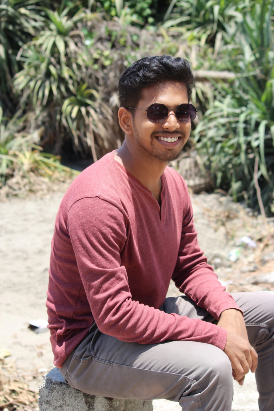 In the image, a young man is seated on a concrete block or stone slab outdoors. He is wearing a red shirt and sunglasses, which adds a pop of color to the scene. The man's face is lit up with a smile, suggesting he is in a good mood. Behind him, there are some bushes and plants that add greenery to the setting.
