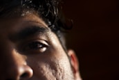 Close-up of a person's face focusing on the left eye and brow. The image captures the texture of the skin with subtle lighting casting shadows, highlighting details like pores and hair.