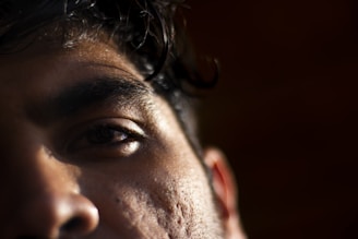 Close-up of a person's face focusing on the left eye and brow. The image captures the texture of the skin with subtle lighting casting shadows, highlighting details like pores and hair.
