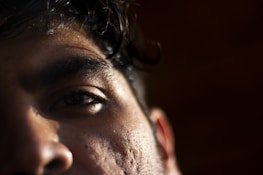 Close-up of a person's face focusing on the left eye and brow. The image captures the texture of the skin with subtle lighting casting shadows, highlighting details like pores and hair.