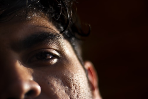 Close-up of a person's face focusing on the left eye and brow. The image captures the texture of the skin with subtle lighting casting shadows, highlighting details like pores and hair.
