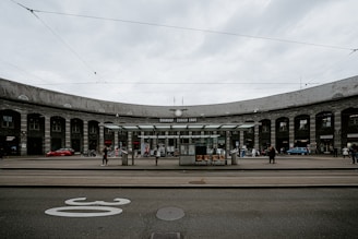 A panoramic view of a train station, with a large, semi-circular stone building in the background labeled 'Bahnhof Zürich Enge'. Several people are walking around or standing near the entrance. The scene includes tram tracks and a tram stop shelter with a few chairs. Overhead, electrical lines for trams are visible against the cloudy sky.