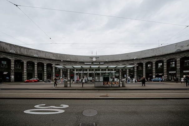 A panoramic view of a train station, with a large, semi-circular stone building in the background labeled 'Bahnhof Zürich Enge'. Several people are walking around or standing near the entrance. The scene includes tram tracks and a tram stop shelter with a few chairs. Overhead, electrical lines for trams are visible against the cloudy sky.
