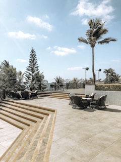 A spacious outdoor patio area with several wicker chairs and tables arranged around it. There are steps leading up to the patio, and it's bordered by a railing. Palm trees and other tropical vegetation surround the patio, with a clear blue sky and a few clouds above.