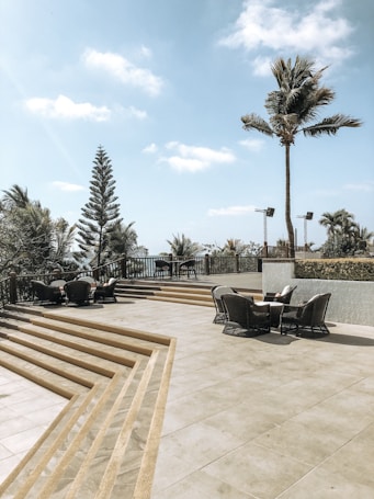 A spacious outdoor patio area with several wicker chairs and tables arranged around it. There are steps leading up to the patio, and it's bordered by a railing. Palm trees and other tropical vegetation surround the patio, with a clear blue sky and a few clouds above.