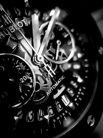 Close-up view of a mechanical watch face featuring exposed gears and intricate details. The watch hands and various mechanical components are intricately designed, showcasing craftsmanship with visible numerals and text indicating 'automatic' and 'Swiss'. The overall appearance is sleek and elegant.