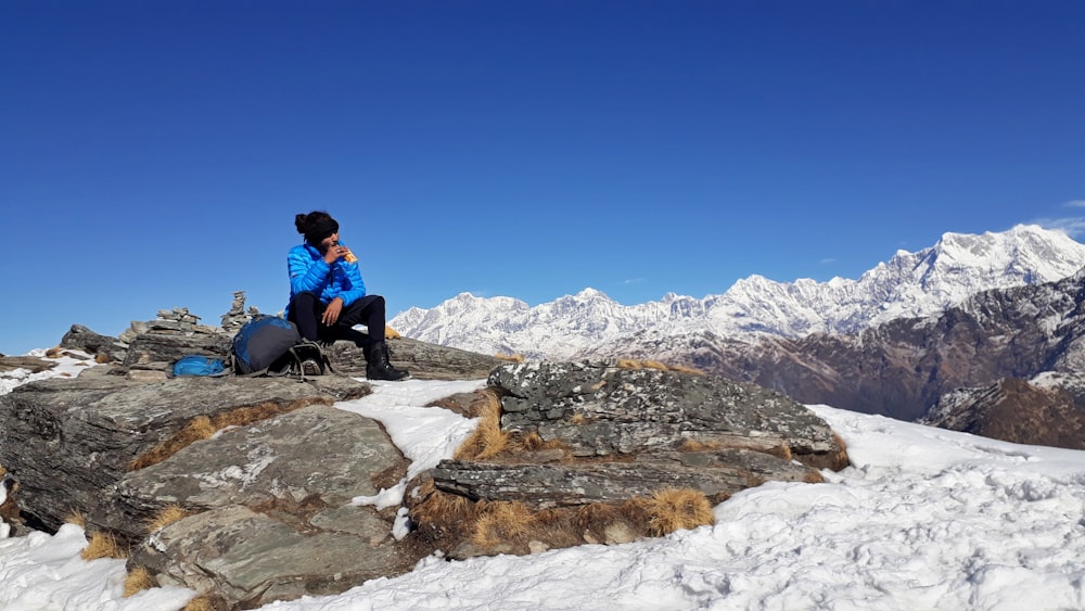 man in blue jacket and black pants sitting on rock on top of hill