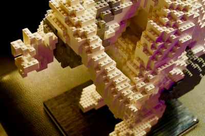 A detailed sculpture constructed from interlocking plastic bricks, featuring intricate layers and an abstract, geometric design. The structure is predominantly composed of white and light gray bricks, with subtle pink and purple hues in certain areas, casting dynamic shadows on a textured black surface below.
