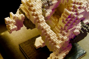 A detailed sculpture constructed from interlocking plastic bricks, featuring intricate layers and an abstract, geometric design. The structure is predominantly composed of white and light gray bricks, with subtle pink and purple hues in certain areas, casting dynamic shadows on a textured black surface below.