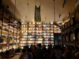 A bustling bar or distillery with shelves stocked with illuminated multicolored bottles filling the entire wall. The room is dimly lit, creating a vibrant and lively atmosphere. Barrels are stacked on one side and a mixed crowd of people are gathered, suggesting a social gathering.
