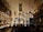 A bustling bar or distillery with shelves stocked with illuminated multicolored bottles filling the entire wall. The room is dimly lit, creating a vibrant and lively atmosphere. Barrels are stacked on one side and a mixed crowd of people are gathered, suggesting a social gathering.