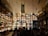 A bustling bar or distillery with shelves stocked with illuminated multicolored bottles filling the entire wall. The room is dimly lit, creating a vibrant and lively atmosphere. Barrels are stacked on one side and a mixed crowd of people are gathered, suggesting a social gathering.