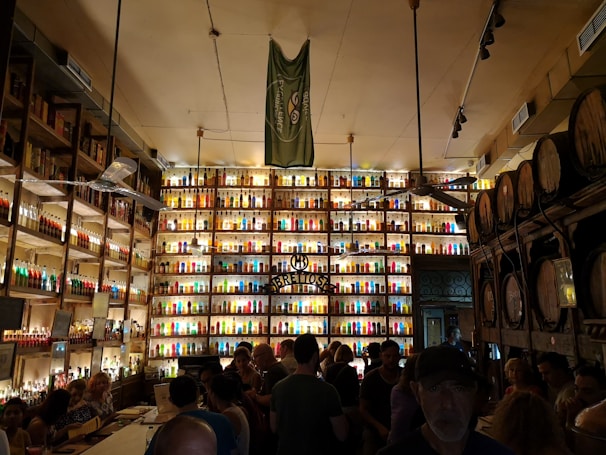 A bustling bar or distillery with shelves stocked with illuminated multicolored bottles filling the entire wall. The room is dimly lit, creating a vibrant and lively atmosphere. Barrels are stacked on one side and a mixed crowd of people are gathered, suggesting a social gathering.