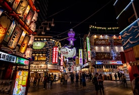 A vibrant and busy street scene at night featuring numerous illuminated signs with Japanese characters. The area is bustling with people walking and exploring, and there's a large, glowing tower in the background. The signs and banners display colorful graphics and artwork, adding to the lively atmosphere.