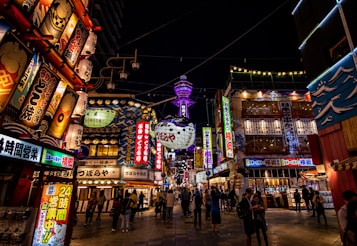 A vibrant and busy street scene at night featuring numerous illuminated signs with Japanese characters. The area is bustling with people walking and exploring, and there's a large, glowing tower in the background. The signs and banners display colorful graphics and artwork, adding to the lively atmosphere.