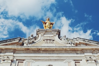 A golden statue of a religious figure with a halo stands atop a classical architectural structure. The structure features ornate sculptures and carvings, with a vivid blue sky and scattered clouds in the background.