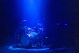 Shaan behind his drum set, illuminated by moody blue stage lighting, lost in the rhythm.