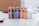 A collection of five bottles of acrylic paint is neatly arranged on a table, each with a distinct color label. The colors are red, black, blue, orange, and beige, with an additional white bottle labeled as acrylic gloss medium. The background appears to be a blurred room interior.