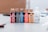 A collection of five bottles of acrylic paint is neatly arranged on a table, each with a distinct color label. The colors are red, black, blue, orange, and beige, with an additional white bottle labeled as acrylic gloss medium. The background appears to be a blurred room interior.