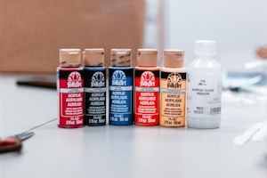A collection of five bottles of acrylic paint is neatly arranged on a table, each with a distinct color label. The colors are red, black, blue, orange, and beige, with an additional white bottle labeled as acrylic gloss medium. The background appears to be a blurred room interior.