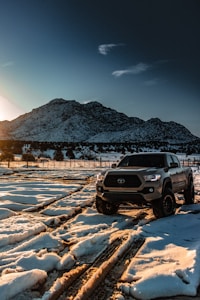 A pickup truck is parked on a snowy landscape with tracks in the snow, set against the backdrop of a large, rugged mountain. The scene is illuminated by the low sun, casting a warm glow across the snow and mountains. Sparse trees are scattered around the area, with a fence running alongside the snowy expanse.