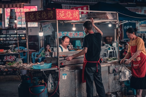 A bustling street food stall with three people engaged in cooking and serving. The scene is illuminated by hanging light bulbs, highlighting skewers and various ingredients. A variety of signs in an Asian script adorn the stall and surrounding shops.
