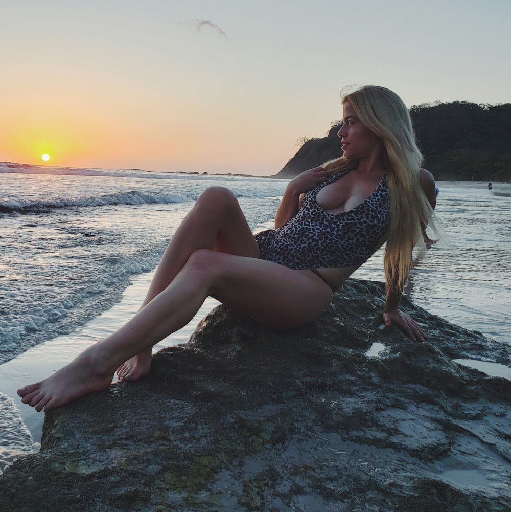 woman wearing black and white bikini sitting on gray rock looking straight on calm sea