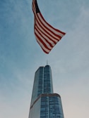A tall skyscraper rises into the sky, made of glass with a sleek modern design. An American flag waves prominently above, contrasting with the blue sky.